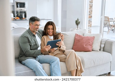 Happy middle aged couple using digital tablet relaxing on couch at home. Smiling mature man and woman holding tab browsing internet on pad device sitting on sofa in living room. Authentic candid photo - Powered by Shutterstock