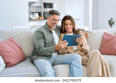 Happy middle aged couple using digital tablet relaxing on couch at home. Smiling mature man and woman holding tab computer browsing internet on pad technology device sitting on sofa in living room. - Powered by Shutterstock