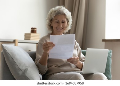 Happy Middle Aged Elderly Woman Sitting On Couch, Holding Computer On Knees Lap, Reading Paper Document Or Mail Letter From Friend. Smiling Mature Old Grandmother Excited By Good News Notification.