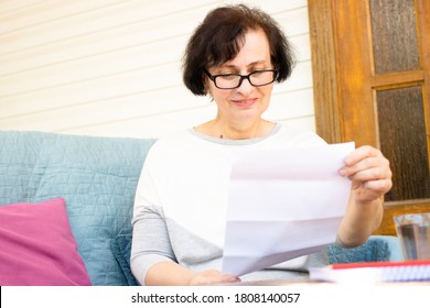 Happy Middle Aged Elderly Woman In Eyeglasses Reading Paper Document Or Postal Letter From Friend, Sitting On Sofa On Home Terrace. Rejection Of Pleasant Paperwork Bank Notice Or Post Correspondence