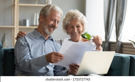 Happy middle aged family couple sitting on couch, looking through paper documents, excited by good banking news. Smiling elderly retired spouses doing paperwork, managing bills together indoors.