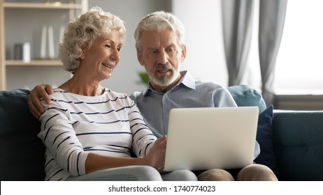 Happy Middle Aged Family Couple Relaxing On Sofa, Using Computer Web Surfing Information Shopping Together At Home. Elderly Man Showing Laptop Apps To Retired Wife, Older Generation With Tech Concept.