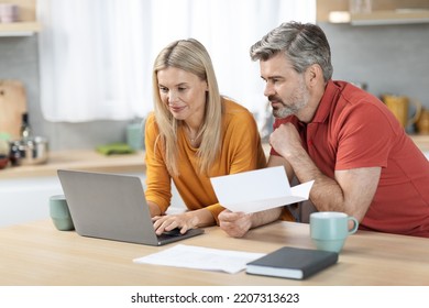 Happy Middle Aged Husband And Wife Sitting At Kitchen Table In Front Of Computer, Holding Papers, Working On Family Budget Together, Paying Bills Online, Copy Space