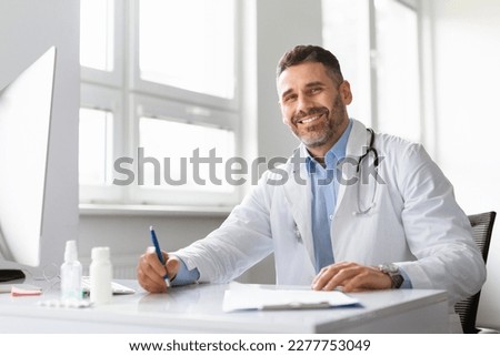 Happy middle aged male doctor in white medical uniform sitting at desk in hospital and smiling at camera, copy space. Excited physician writing in medical journal in clinic. Medicine concept