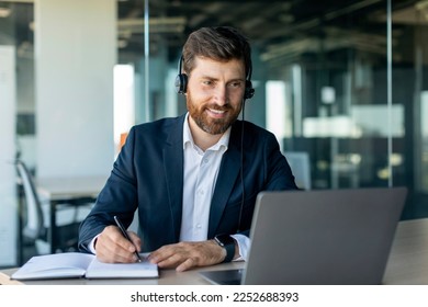 Happy middle aged male manager in headphones looking at laptop and making notes at workplace in office interior. Work, business, study and data analysis remote, client support, ad and offer - Powered by Shutterstock