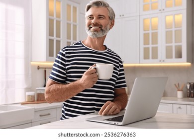 Happy middle aged man with cup of drink and laptop at white marble table in kitchen - Powered by Shutterstock