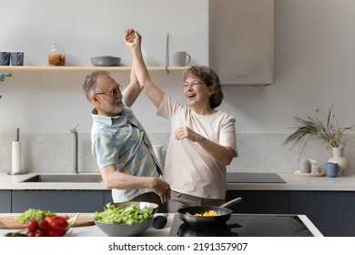 Happy Middle Aged Man Having Fun With Laughing Elderly Senior Wife, Dancing To Disco Music In Modern Kitchen, Distracted From Preparing Healthy Food, Weekend Domestic Family Couple Hobby Activity.