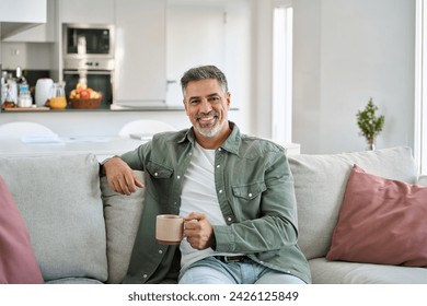 Happy middle aged man holding coffee cup relaxing on couch at home. Smiling mature older man drinking tea looking at camera sitting on cozy sofa chilling in modern kitchen living room. Portrait. - Powered by Shutterstock