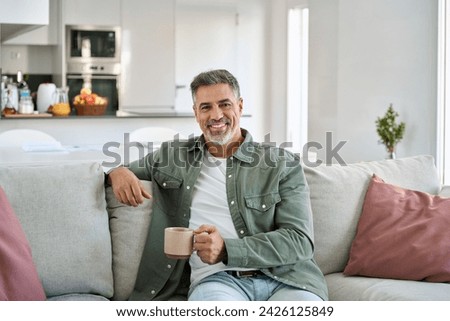 Happy middle aged man holding coffee cup relaxing on couch at home. Smiling mature older man drinking tea looking at camera sitting on cozy sofa chilling in modern kitchen living room. Portrait.