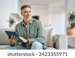 Happy middle aged man using digital tablet relaxing on couch at home. Mature male user holding tab computer holding pad technology device sitting on sofa in living room looking away. Copy space.