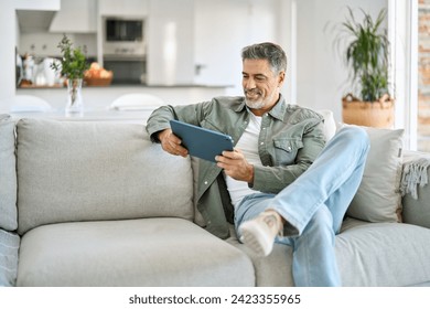 Happy middle aged man using digital tablet relaxing on couch at home. Happy mature male user holding tab computer browsing internet on pad technology device sitting on sofa in living room. - Powered by Shutterstock