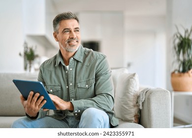 Happy middle aged man using digital tablet relaxing on couch at home. Mature male user holding tab computer holding pad technology device sitting on sofa in living room looking away. Copy space.