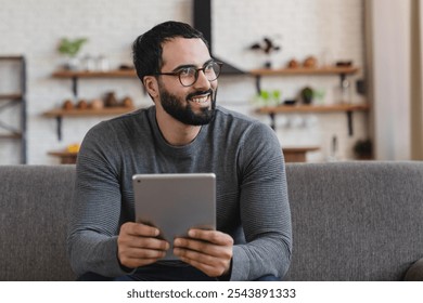 Happy middle aged man using digital tablet relaxing on couch at home. Adult male user holding tab computer holding pad technology device sitting on sofa in living room looking away. Copy space. - Powered by Shutterstock