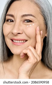 Happy middle aged mature asian woman, senior older 50 year lady looking at camera touching her face applying eye antiaging anti wrinkle skin care cream, isolated on white close up face portrait.