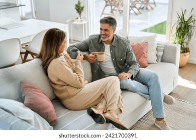 Happy middle aged mature couple talking drinking coffee relaxing on couch at home. Relaxed older man and woman in love sitting on sofa enjoying conversation in living room. Candid photo. - Powered by Shutterstock