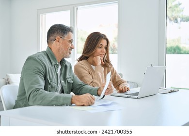 Happy middle aged mature man and woman paying bills online at home. Older senior couple using laptop computer checking insurance or financial invoice counting taxes sitting at table in living room. - Powered by Shutterstock