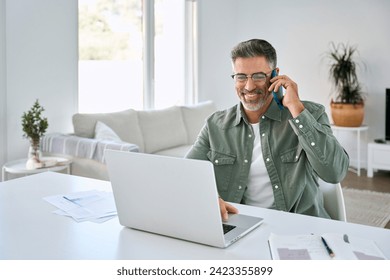 Happy middle aged mature man senior entrepreneur wearing eyeglasses talking on mobile phone looking at laptop using computer calling customer service support on smartphone working at home office.