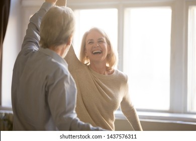 Happy middle aged mature woman enjoying dancing with elder husband at home, active healthy senior old couple man and woman pensioners having fun in waltz laughing bonding celebrating anniversary