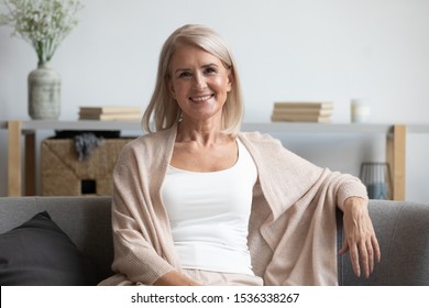 Happy Middle Aged Mature Woman Looking At Camera Sitting Alone On Sofa At Home, Smiling Attractive Senior Adult Old Retired Lady Relaxing Sitting On Couch In Apartment Living Room Posing For Portrait