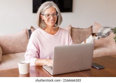 Happy Middle Aged Mature Woman Using Wireless Laptop Apps Browsing Internet Sit At The Table, Smiling Gray-haired Lady Working Distantly On Computer, Communicating Online Looking At Screen At Home