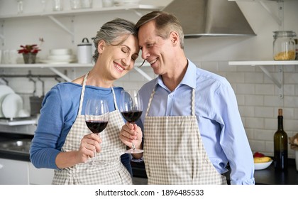 Happy Middle Aged Old Senior Family Couple In Love Wearing Apron Holding Glasses, Drinking Wine Standing In Kitchen, Enjoying Bonding While Cooking At Home, Celebrating Anniversary, Valentines Day.