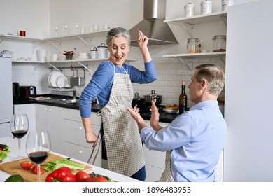 Happy Middle Aged Older 50s Family Couple Having Fun Cooking Together. Smiling Active Senior Wife Dancing To Music While Husband Clapping Hands Enjoying Funny Dance, Celebrating At Home In Kitchen.