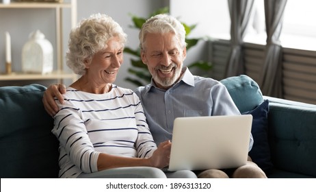 Happy Middle Aged Older Retired Family Couple Using Computer Applications, Having Fun Web Surfing Internet, Shopping Online Or Communicating Distantly Relaxing Together On Cozy Sofa In Living Room.