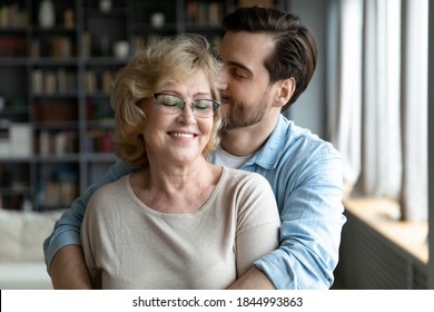 Happy Middle Aged Older Woman In Eyeglasses Standing With Closed Eyes, Enjoying Tender Loving Moment With Grown Son At Home. Devoted Young Man Showing Care To Smiling Retired Old Mother Indoors.