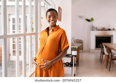 Happy middle aged pregnant woman with big tummy relaxing at home. Portrait of mature pregnant african american woman standing near the window and caressing her belly. Mid black lady expecting child. - Powered by Shutterstock