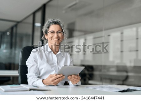 Happy middle aged professional business woman using tab computer in office. Mature female bank manager, smiling older executive holding digital tablet looking at camera sitting at work desk. Portrait.