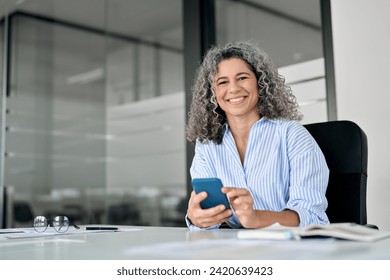 Happy middle aged senior curly business woman holding phone in office looking at camera. Smiling mature female entrepreneur, older professional worker using cellphone working at desk, portrait. - Powered by Shutterstock