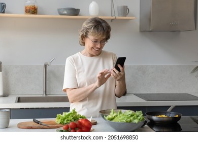 Happy middle aged senior retired woman in eyeglasses using smartphone, communicating online in social network or searching interesting recipes preparing healthy food in modern kitchen on weekend. - Powered by Shutterstock