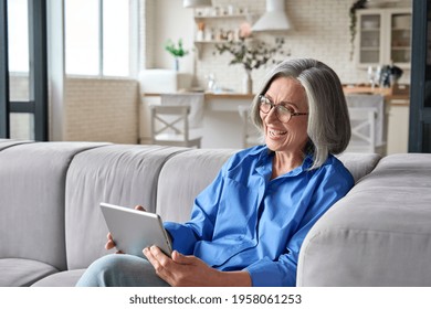 Happy middle aged senior woman sitting on couch holding using digital tablet device during video call with family friends. Older female reading ebook or watching zoom video meeting online.