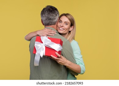 Happy Middle Aged Spouses Hugging On Yellow Background, Woman Holding Gift Box And Smiling At Camera, Couple Celebrating Anniversary Together