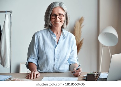 Happy middle aged stylish woman fashion designer wears glasses standing at workplace desk, portrait. Smiling sophisticated 60s old lady dressmaker drawing sketches, looking at camera in modern atelier