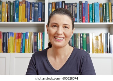 Happy Middle Aged Woman With Blue T-shirt In Front Of Lots Of Books, Having An Live Video Chat With Her Family Or Onlne Dating