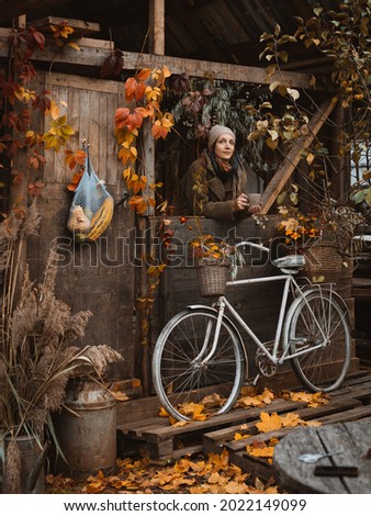 Happy middle aged woman with cup of coffee on rustic terrace of old wooden house enjoying morning in village on autumn day, vintage bicycle leaning on wall and seasonal vegetables hanging on door