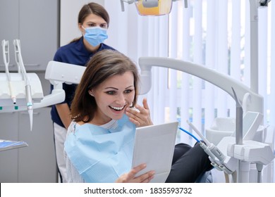 Happy Middle Aged Woman With Doctor Dentist Looking In Mirror At Teeth, Sitting In Dental Chair. Medicine, Dentistry And Health Care Concept