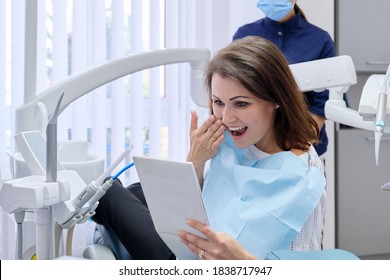 Happy Middle Aged Woman With Doctor Dentist Looking In Mirror At Teeth, Sitting In Dental Chair. Medicine, Dentistry And Health Care Concept