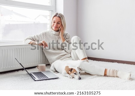 Happy middle aged woman with dog using her laptop and mobile phone at cozy white home. Female freelancer working in living room
