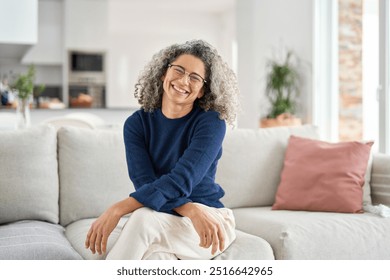 Happy middle aged woman in her 50s sitting on sofa in living room at home. Smiling beautiful mature older lady with curly gray hair relaxing on couch looking at camera on couch in cozy house. Portrait - Powered by Shutterstock