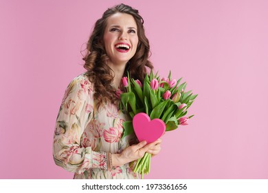 happy middle aged woman with long wavy brunette hair with tulips bouquet and pink heart isolated on pink background.