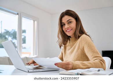 Happy middle aged woman looking at camera holding documents hybrid working at home table. Mature lady paying bills online checking financial papers invoice making baking payments using laptop computer - Powered by Shutterstock