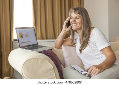 Happy Middle Aged Woman Looking At Her Finances On Computer Screen And Talking On A Phone