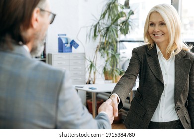 Happy middle aged woman shakes hands with handsome businessman after successful negotiations.Elegant female in stylish formal suit on interview in modern office, smiling, happy about getting a new job