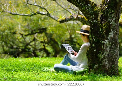 Happy Middle Aged Woman Sitting On Green Grass Using Tablet Computer In The Park