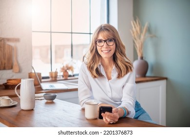 Happy middle aged woman standing in the kitchen at home and text messaging.  - Shutterstock ID 2201376873