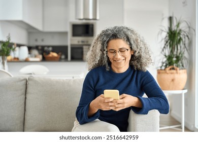 Happy middle aged woman using smartphone sitting on couch at home. Mature lady wearing glasses holding cellphone browsing internet, texting messages on mobile cell phone technology on sofa. - Powered by Shutterstock