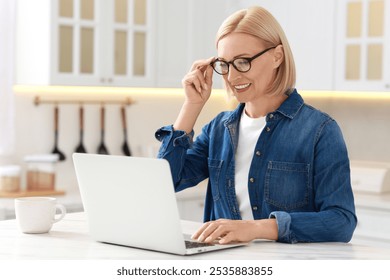 Happy middle aged woman using laptop at white marble table in kitchen - Powered by Shutterstock