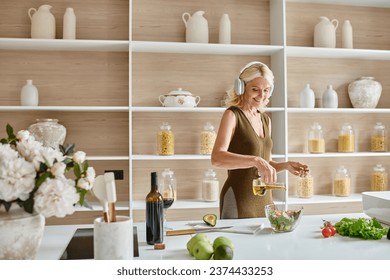 happy middle aged woman in wireless headphones making salad near bottle and glass of red wine - Powered by Shutterstock